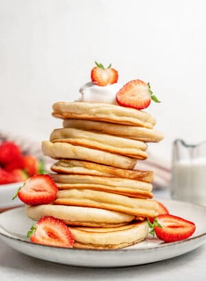A tall stack of gluten-free pikelets on a plate garnished with whipped cream and sliced strawberries.